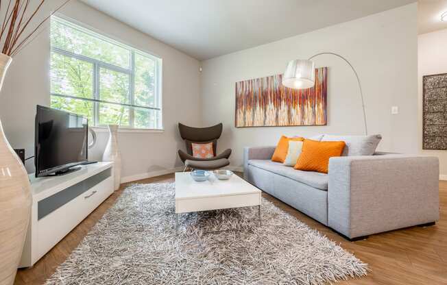 Model home living room with light grey couch, lamp that hangs over the couch, windows and chair in the corner.  Flooring is wood with an area rug in the center