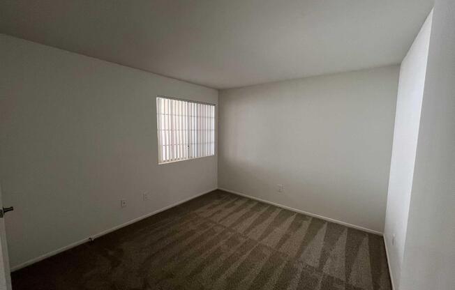 A small, empty room with plain white walls and beige carpet. There's a barred window on the left, allowing natural light to filter in. The space is unadorned and minimalistic, giving it a clean and airy feel, suitable for various uses.