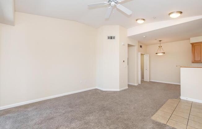 An empty living space with light beige walls and plush, neutral carpet. A ceiling fan is visible in the upper left corner, and a small kitchen area with wooden cabinetry is in the background. The room is well-lit with natural light and features a tile section near the kitchen.
