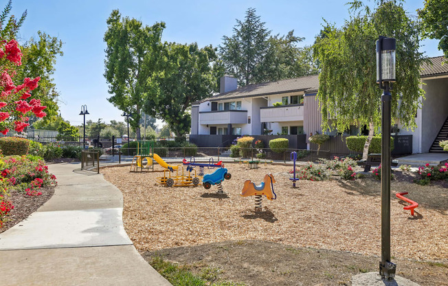 A playground with a slide, swings, and a climbing frame is surrounded by trees and flowers.