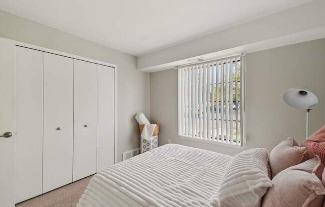 A bedroom with a bed, pillows, a lamp, and a window with blinds.