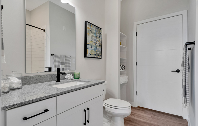 A modern bathroom featuring a white vanity with a gray countertop, a black faucet, and a mirror above. There's a shower with white tiles in the background, a white door, and open shelving displaying decorative items. The floor has a wood-like appearance, creating a warm ambiance.
