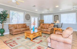 A cozy living room featuring two brown leather sofas, a beige armchair, and a wooden coffee table. A vase with flowers sits on the table. The room has light-colored walls, tiled flooring, and large windows with white blinds, providing natural light. A small table and chairs are visible in the background.