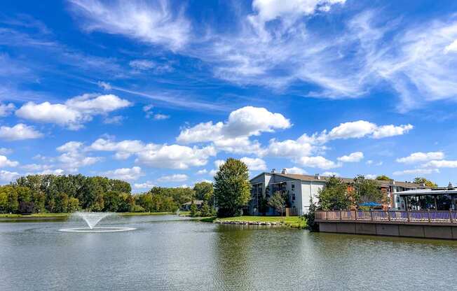A serene lake with a fountain in the middle, surrounded by trees and buildings under a clear blue sky.
