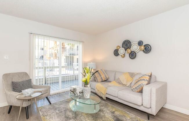 A modern living room with a light gray sofa adorned with yellow and patterned throw pillows. In front, there's a glass coffee table with decorative items. A patterned rug lies on the floor. To the side, a cozy chair complements the space, and large windows let in natural light, showing a glimpse of an outdoor balcony.