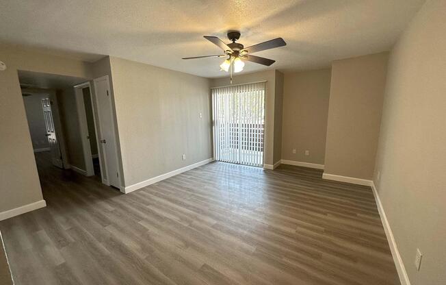 A spacious, empty room with light-colored walls and wood-like flooring, featuring a ceiling fan. There is a sliding glass door leading to an outdoor area with blinds partially drawn. The room is well-lit with natural light, giving it a bright and airy feel. Doorways lead to other areas.