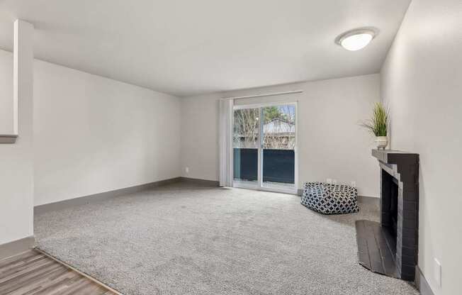 A living room with a grey carpet, a fireplace, and a window with a view of trees.