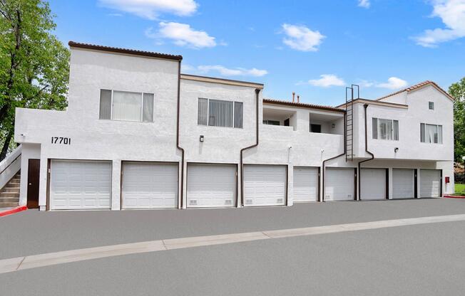 A modern two-story white building with multiple garage doors on the ground floor, situated in a landscaped area under a clear blue sky. The design features a mix of windows and balconies, enhancing its contemporary aesthetic.