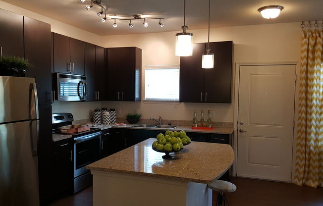 Photo of kitchen with large island, granite counter-tops, modern cabinetry, microwave, electric stove, dishwasher, refrigerator, and double sided sink, from a different angle, showing it next to the entry door.