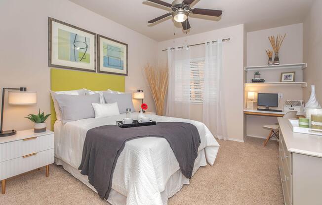 Cozy bedroom featuring a queen-sized bed with gray and white bedding, decorative pillows, and a bright green headboard. A ceiling fan and two lamps provide lighting, while a small desk and chair, along with decorative plants and framed artwork, enhance the inviting atmosphere. Natural light filters through sheer curtains.