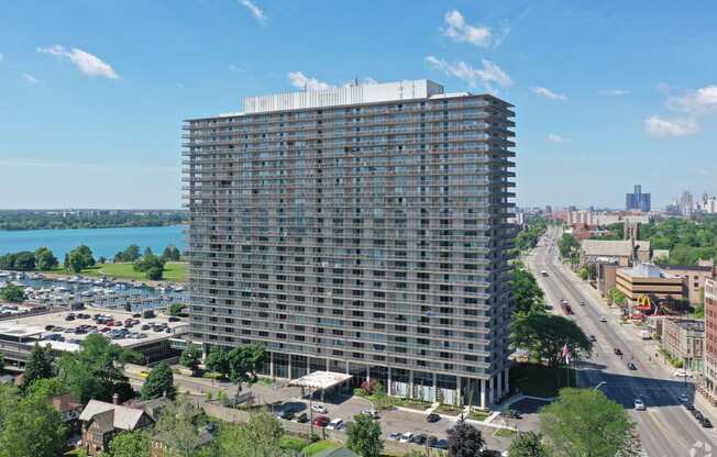 Exterior of The Jefferson. The building stands 30 stories tall. The building is made of concrete and glass. There are green trees and blue skies. You can see the Detroit River in the background. The building is located on Jefferson Ave.