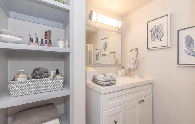 A modern bathroom featuring a white vanity with a mirror and overhead lighting. Shelves on the left hold neatly arranged towels, nail polish, and bath accessories. Decorative wall art with coral designs is framed on the walls, and a serene color palette creates a calming atmosphere.