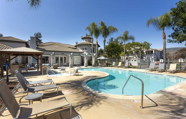 A sunny pool area featuring a curved swimming pool surrounded by lounge chairs and palm trees. In the background, a landscaped building with a tiled roof is visible, along with shade structures and umbrellas, creating a relaxing outdoor atmosphere.