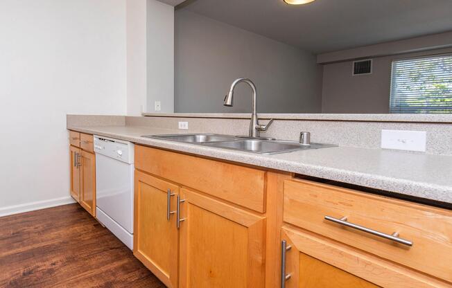 Modern kitchen featuring light wood cabinetry, a double sink, and a built-in dishwasher. The countertop is gray speckled, and the space has a neutral color scheme. A window in the background allows natural light to enter.