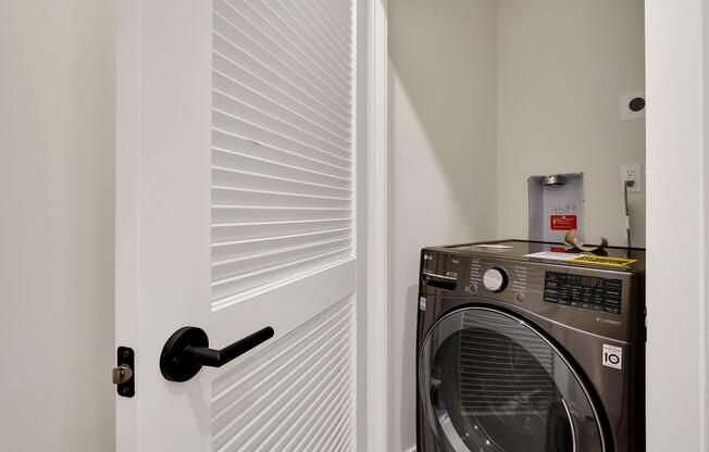 A modern laundry room featuring a front-loading washing machine and a partially open door with contemporary slatted design. The space is well-lit and includes electrical outlets and labels on the washing machine for instructions or settings.