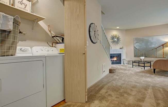 A convenient laundry area here at Aspen Creek featuring a full-size washer and dryer tucked into a dedicated closet with shelving above, opening into a carpeted living space with a staircase, neutral walls, and a cozy fireplace accented by a mantle, decorative mirror, and artwork, creating a functional layout that blends everyday convenience with comfortable living.