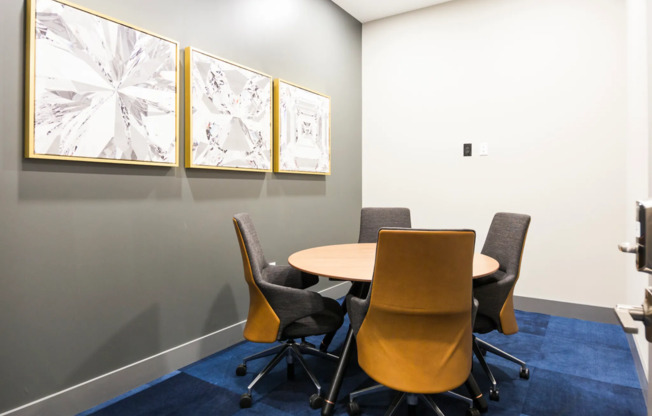 A private meeting area with four rolling chairs around a table, bright blue carpeting, and abstract art on the walls.