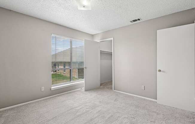 A room with a carpeted floor, a window with blinds, and a view of a fence and a building outside.