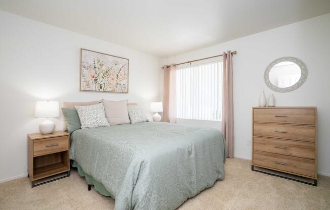 A bright and inviting bedroom featuring a queen-sized bed with a light green blanket, complemented by decorative pillows. There are two bedside lamps, a wooden dresser, and a round mirror on the wall. A window with sheer curtains allows natural light to enter, enhancing the cozy atmosphere.