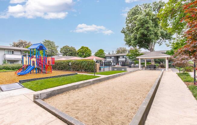Playground area featuring a colorful play structure with slides and climbing features, surrounded by a grassy area. Adjacent is a bocce ball court made of sand, lined with concrete. There are shaded seating areas and well-maintained landscaping, creating a pleasant outdoor recreational space.