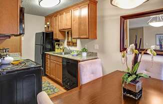 A cozy kitchen here at Whispering Cedars with light brown cabinets, granite countertops, and black appliances, featuring a stainless steel sink, dishwasher, and adjacent dining area with a wooden table and decorative mirror, creating a warm, inviting feel.