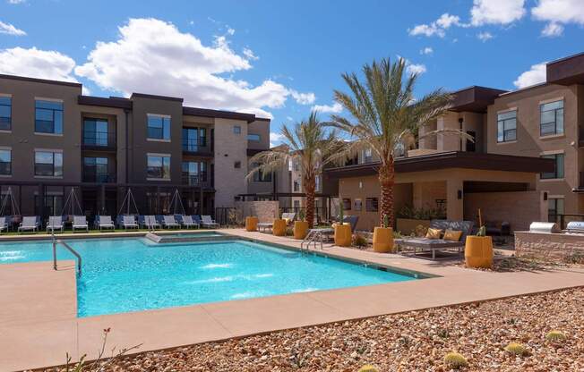 A swimming pool is surrounded by chairs and palm trees in front of a building.