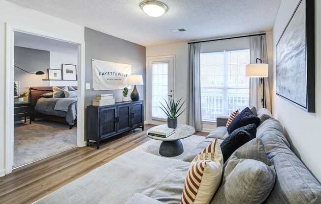 A living room with a grey sofa and a bedroom with a bed and a nightstand in the background.