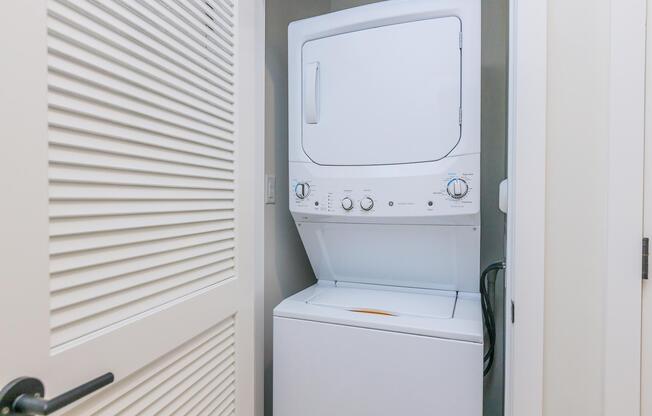 A compact laundry unit with a stacked washer and dryer, located in a small laundry space that features a door with horizontal slats. The appliances are white, and the area has neutral-colored walls.