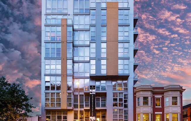 A modern building with a glass facade is illuminated from within, with cars parked in front and a street scene in the foreground.