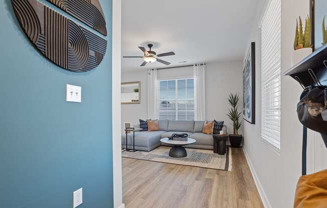 looking from a hallway into a large living room with large windows, an illuminated fan, and model furnishings