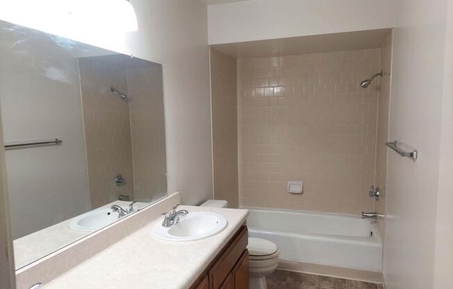 A clean, modern bathroom featuring a bathtub with a showerhead, a large mirror above a dual sink vanity, and beige tiled walls. The floor is light-colored, and a towel bar is mounted next to the mirror. The overall decor is neutral and simple, providing a spacious feel.