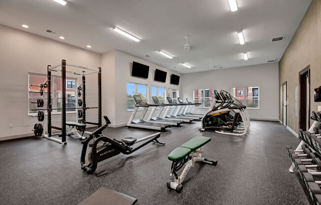 A bright fitness center here at The Park featuring modern equipment, rubber flooring, large windows, multiple treadmills, weight machines, benches, and mounted TVs, creating an energetic and inviting workout space.
