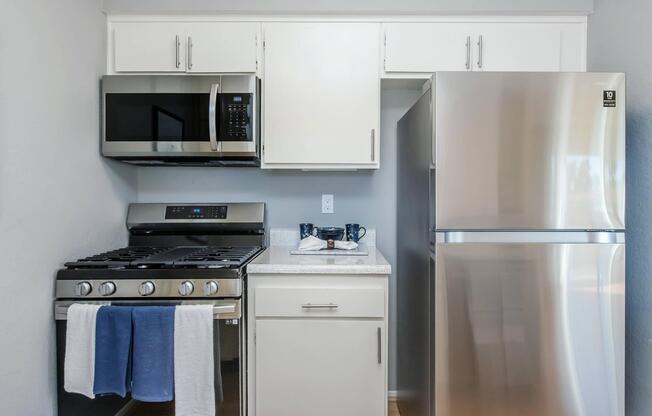 Modern kitchen featuring stainless steel appliances including a refrigerator, oven with a stovetop, and microwave. The cabinets are white, and there is a light-colored countertop with a small tray and dish on it. Clean and organized design, with a blue towel hanging from the oven.