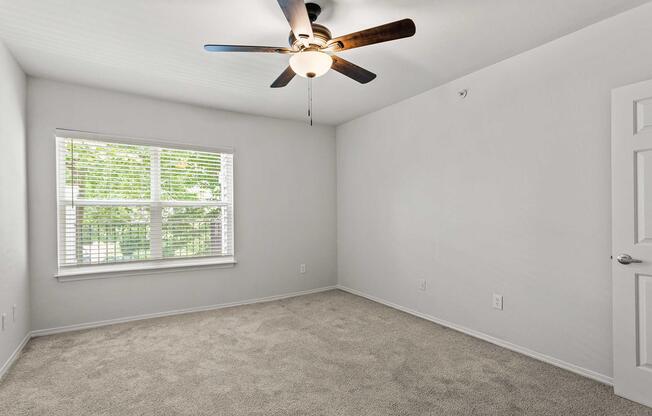A bright, empty room featuring light-colored walls and plush carpet. A ceiling fan hangs from the ceiling, and a window with white blinds allows natural light to enter, illuminating the space. The room is unadorned and ready for personalization.