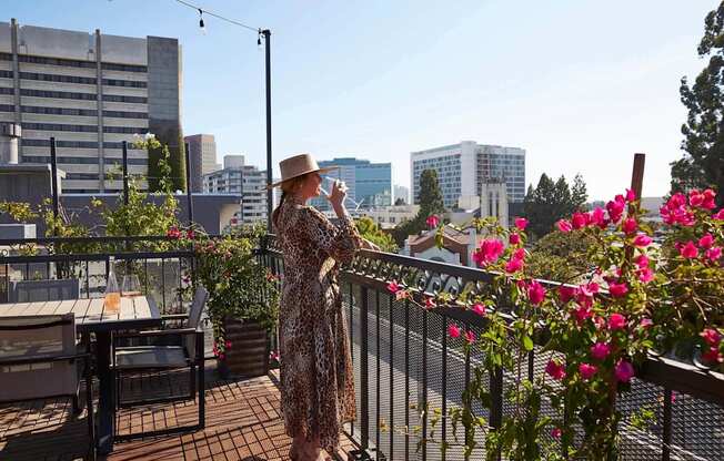 A woman in a leopard print dress and hat stands on a balcony with pink flowers.