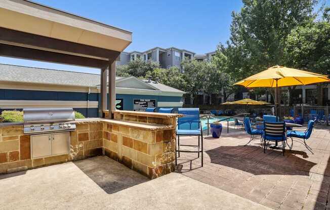 A grilling area is on a patio with blue chairs and yellow umbrellas with the pool in the background at Encore at Buckingham Apartments in Richardson, TX