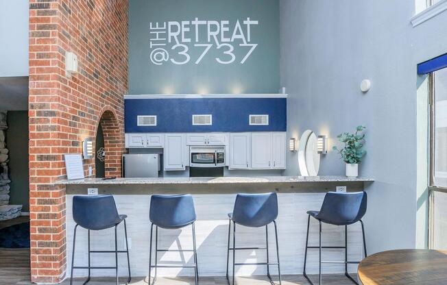Modern interior of a stylish kitchen bar area featuring four black high stools. The backdrop includes a large wall with the text "THE RETREAT @3737" in bold lettering. The kitchen has white cabinets, stainless steel appliances, and a sleek countertop, with natural light coming through large windows.