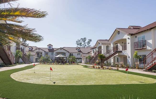 A golf hole with a red flag on a sunny day in front of a row of houses.