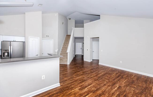 Spacious and modern interior of a residential living area featuring hardwood floors, a staircase leading to an upper level, and an open kitchen with stainless steel appliances. The walls are painted in neutral tones, creating a bright and inviting atmosphere.