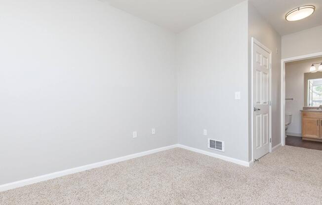 Empty room with soft beige carpet and light gray walls. A door is on the left side leading to a hallway, and there is a bathroom visible in the background. Natural light from a window illuminates the space, creating a clean and minimalistic aesthetic.
