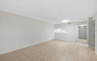 Empty room with beige tile flooring and neutral-colored walls. Features include a light fixture on the ceiling, a small opening to a kitchen area, and a doorway leading to a hallway. Natural light floods the space, creating a bright and open atmosphere.