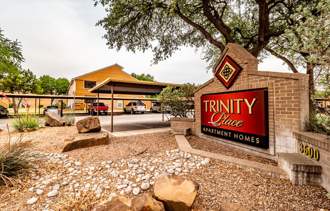 A landscaped community entrance here at Trinity Place featuring a brick monument sign displaying the property name. Desert-style ground cover, decorative rocks, and mature trees surround the sign, while covered parking and apartment buildings are visible in the background, creating a welcoming first impression of the apartment community.