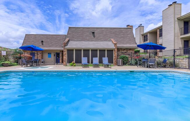 A clear blue swimming pool in front of a single-story building with a sloped roof. The pool area features lounge chairs and umbrellas, surrounded by well-maintained landscaping. In the background, additional buildings are visible under a partly cloudy sky.