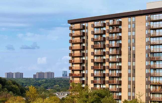 A tall building with many windows is in the foreground with trees and other buildings in the background.