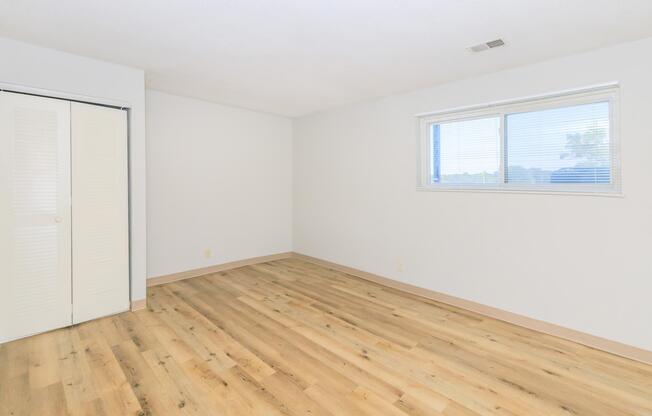 A bright, empty room with light-colored walls and a large window. The floor is wooden with a light finish. There's a closet with sliding doors on one side of the room. The space is well-lit, suggesting a clean and inviting atmosphere, perfect for customization or personal decoration.