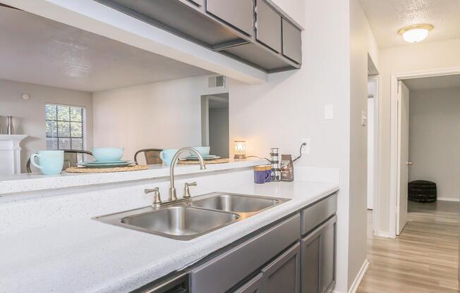 A modern kitchen featuring a double sink, dark cabinets, and a countertop. In the background, there is a dining area with a table set with plates and cups. Natural light enters through a window, and a doorway leads to another room. The overall decor is clean and contemporary.