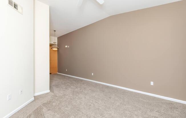 A spacious, empty room with beige walls and light-colored carpet. The ceiling features a ceiling fan, and there is a doorway leading to another space. The overall atmosphere is bright and open, ideal for decorating or furnishing.
