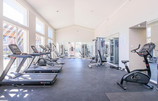 A bright and spacious fitness center featuring various exercise equipment, including treadmills, stationary bikes, and weight machines. Large windows provide ample natural light, and mirrors line the walls, enhancing the open feel of the gym space.