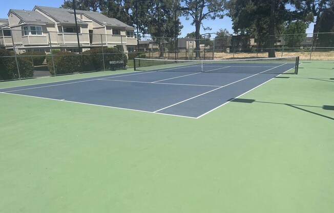 A tennis court with dark blue markings on a green surface. Surrounding the court are trees and residential buildings in the background. The area appears well-maintained and is bathed in daylight, indicating clear weather.