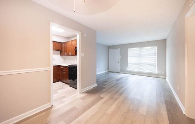 A room with a kitchen area in the background and a large window with blinds.